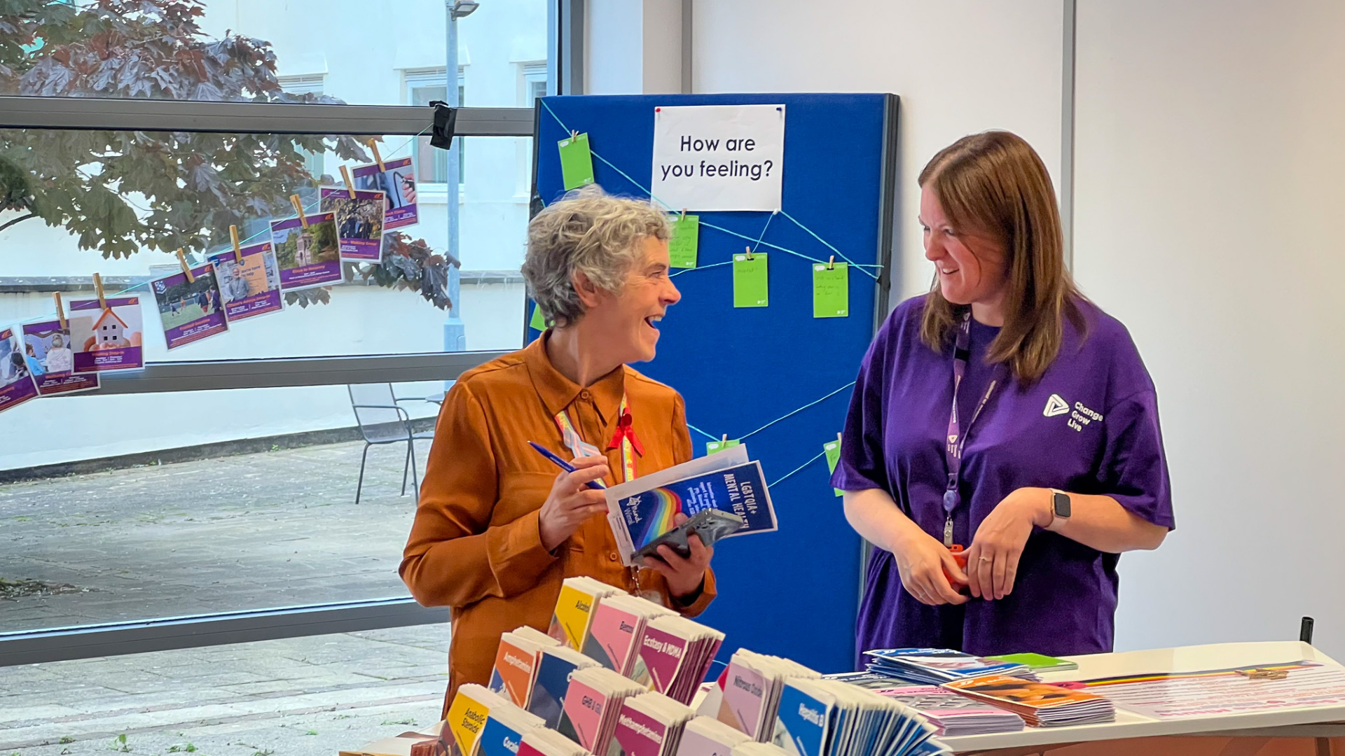 Kath from Sahir and Denise at a wellbeing information stand (16:9) Kath from Sahir and Denise from Wirral Ways smiling and talking at an information stand, surrounded by colourful leaflets and a "How are you feeling?" activity board in a light-filled room.