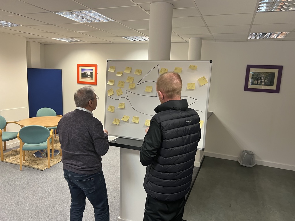 SU Forum - Gerry Pangallis and Service User looking at board Two people participating in a service user forum, standing in front of a whiteboard with a boat outline covered in yellow sticky notes. Each note appears to share thoughts, ideas, or feedback. The room has a calm, informal setting with round tables and framed artwork on the walls.