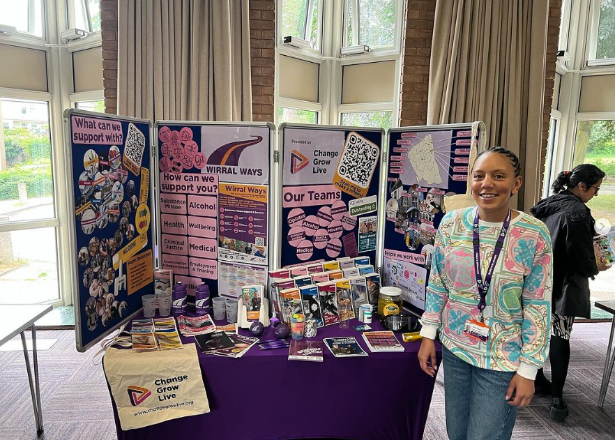Becky at Wirral Ways Community Stall Becky from Wirral Ways standing at a vibrant community event stall, smiling in front of a display filled with leaflets, promotional items, and posters. The stand highlights support available from Wirral Ways, covering areas like substance misuse, health, wellbeing, and criminal justice. The event is set in a bright, welcoming venue with large windows and natural light.